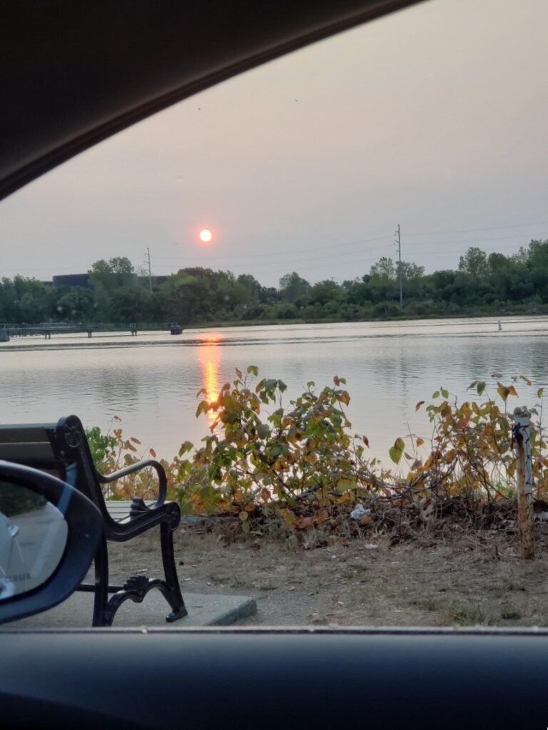 Photo of the sun shining over a park bench that is facing towards the water